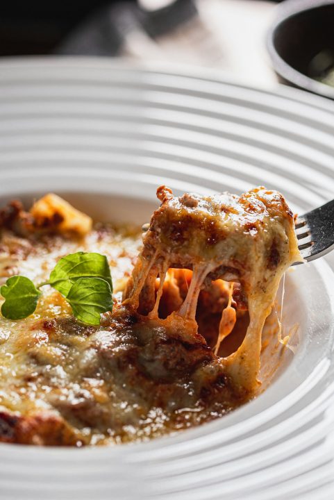 Fork lifting a cheesy bite of lasagna from a white bowl