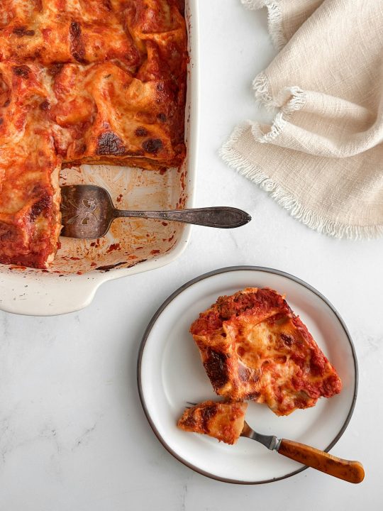 A slice of lasagna served from a white baking dish onto a white plate with a vintage fork