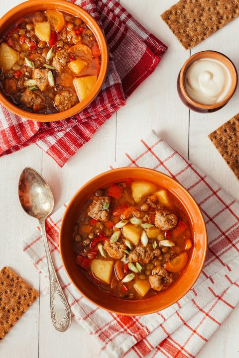 Two bowls of lentil soup with meatballs on a table with crackers