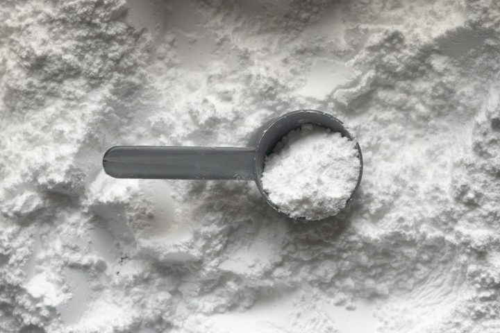 A grey plastic scoop filled with white protein powder resting on a pile of powder