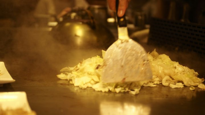 A metal spatula stirring chopped cabbage on a hot griddle