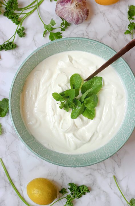 A bowl of yogurt with a spoon in it, with fresh herbs and garlic nearby
